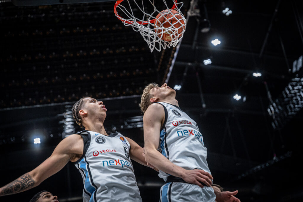 Zwei Basketballspieler in weißen Trikots springen unter einem Korb in einer Halle mit dunkler Decke und hellen Deckenleuchten.