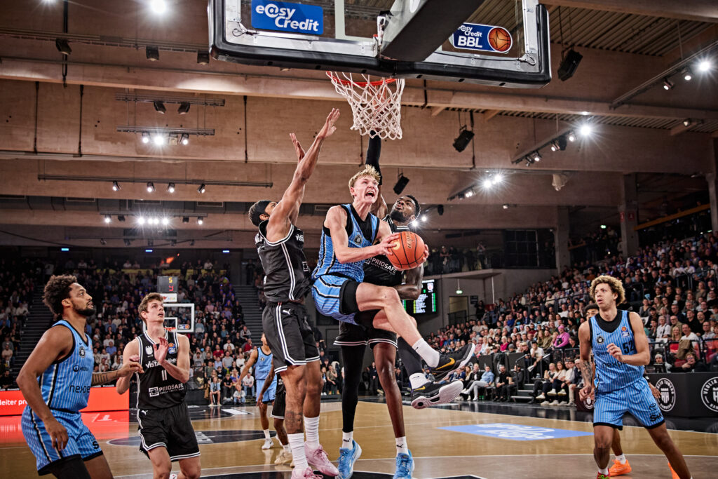 Basketballspieler in blauen Trikots versuchen, gegen Verteidiger in schwarzen Trikots einen Korb zu erzielen, Zuschauer im Hintergrund.