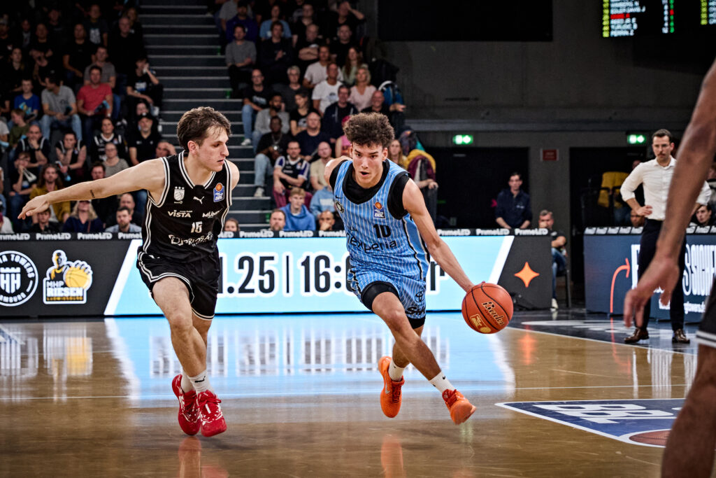 Zwei Basketballspieler in Aktion auf dem Spielfeld, einer in blau-schwarzem Trikot mit Ball, der andere in schwarzem Trikot verteidigt.