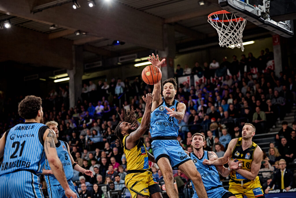 Mehrere Basketballspieler in blauen und gelben Trikots kämpfen um den Ball unter dem Korb in einer vollen Halle.