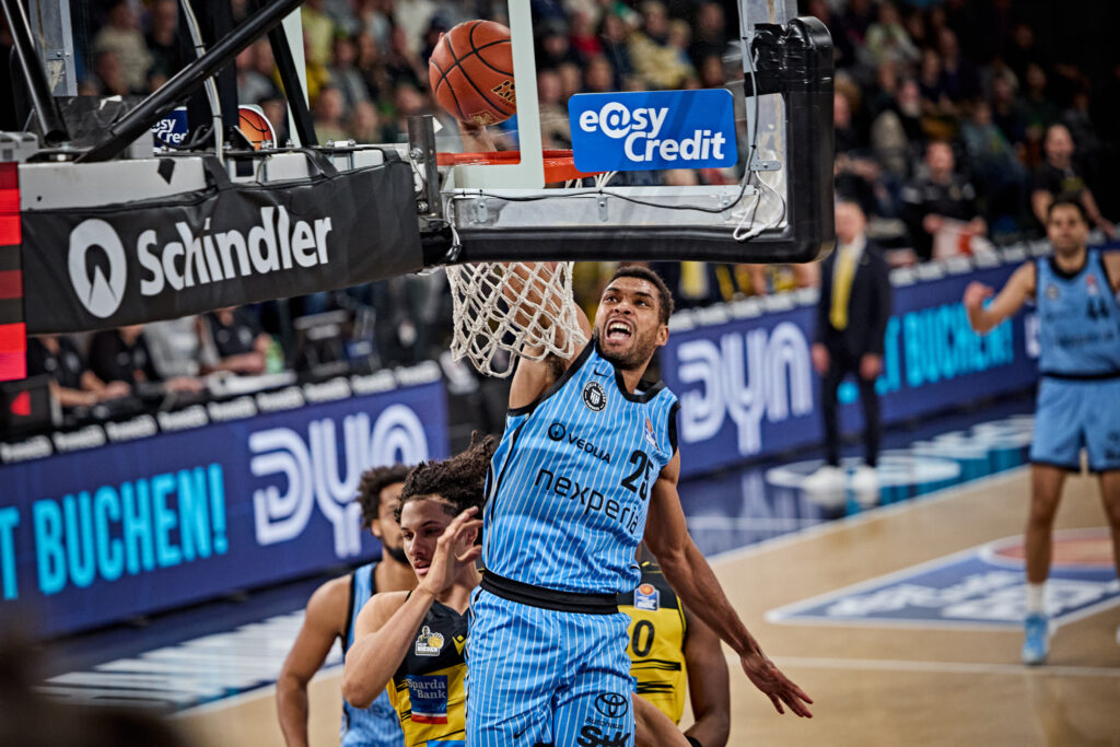 Basketballspieler in blauem Trikot mit der Nummer 21 springt zum Korb, um den Ball zu werfen, während ein Spieler in gelbem Trikot verteidigt.