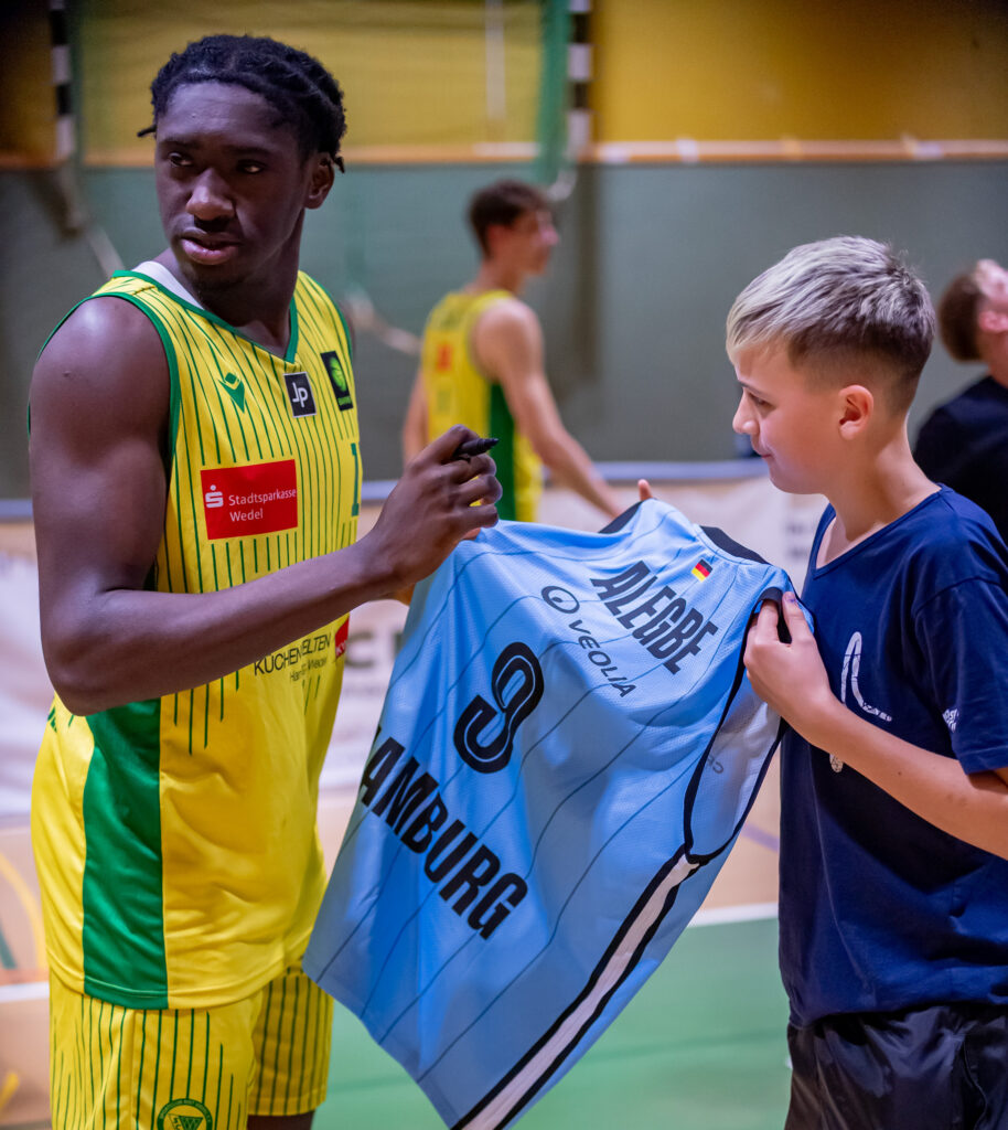 Basketballspieler in gelb-grünem Trikot übergibt blaues Trikot mit der Nummer 9 an Jungen in blauem T-Shirt in einer Sporthalle.