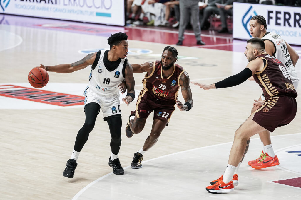 Basketballspieler in Aktion auf dem Spielfeld, einer in weißem Trikot mit Ball, mehrere in dunkelrotem Trikot in Verteidigungsposition.