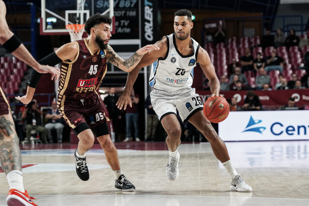 Zwei Basketballspieler in Aktion, einer in weißem Trikot mit Ball, der andere in dunkelrotem Trikot verteidigt.