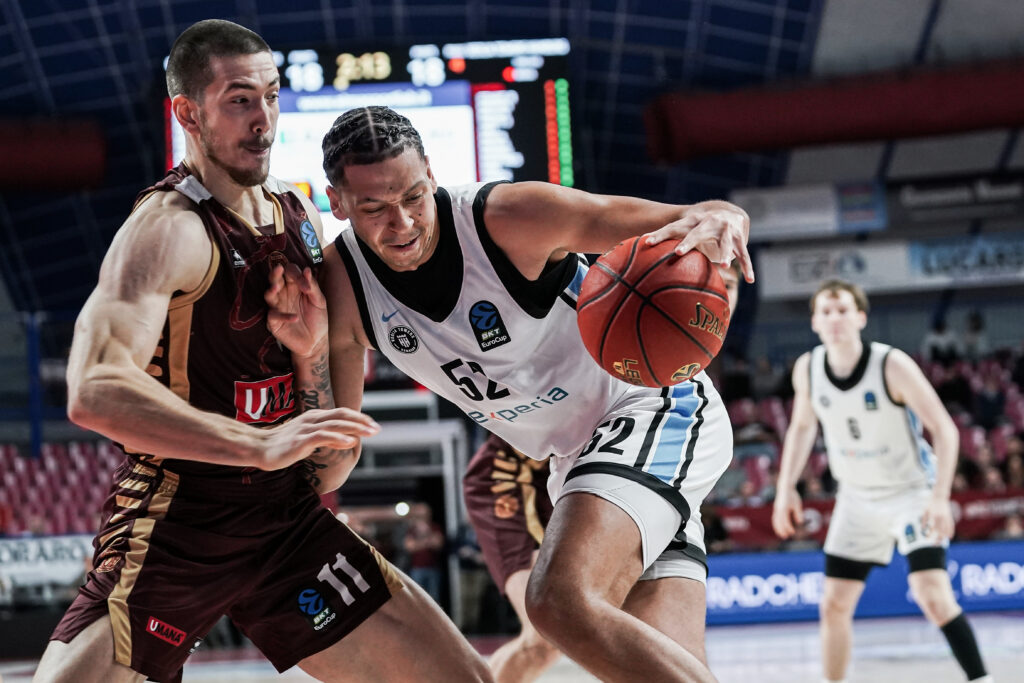 Zwei Basketballspieler in Aktion, einer in weißem Trikot mit Ball, der andere in braunem Trikot verteidigt, im Hintergrund weitere Spieler und Anzeigetafel.