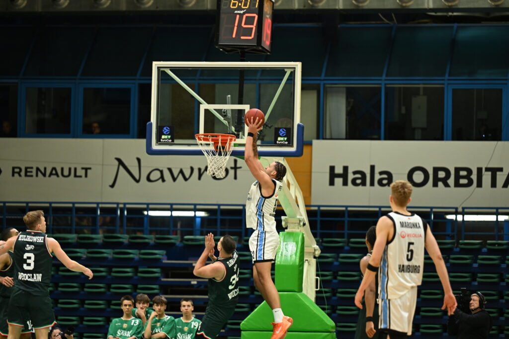 Basketballspieler springt zum Korb, um den Ball zu werfen, umgeben von mehreren Gegenspielern in einer Halle.