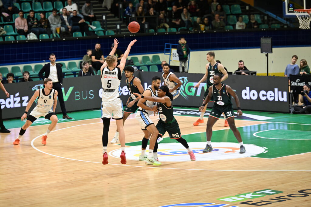 Basketballspieler in Aktion auf dem Spielfeld, ein Spieler in weißem Trikot wirft den Ball, mehrere Spieler in schwarzen Trikots verteidigen.