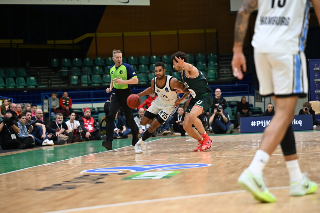 Zwei Basketballspieler, einer in weißem Trikot mit Ball, der andere in schwarzem Trikot verteidigt, Schiedsrichter und Zuschauer im Hintergrund.