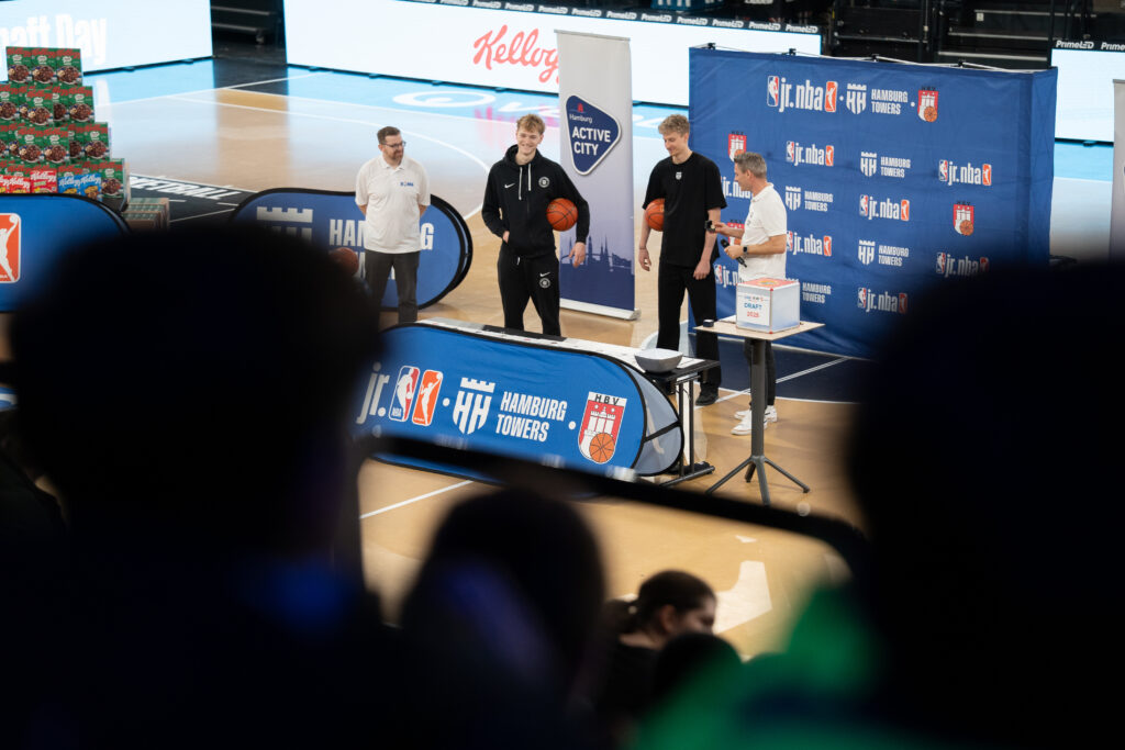 Drei junge Männer stehen in einer Sporthalle auf einem Basketballfeld, zwei halten Basketballs, im Hintergrund Banner mit Logos von jr.nba und Hamburg Towers.