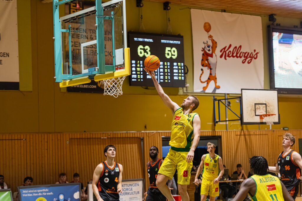 Basketballspieler in gelbem Trikot springt zum Korb, um den Ball zu werfen, andere Spieler beobachten das Spiel in einer Halle mit Anzeigetafel und Kellogg's-Werbung.