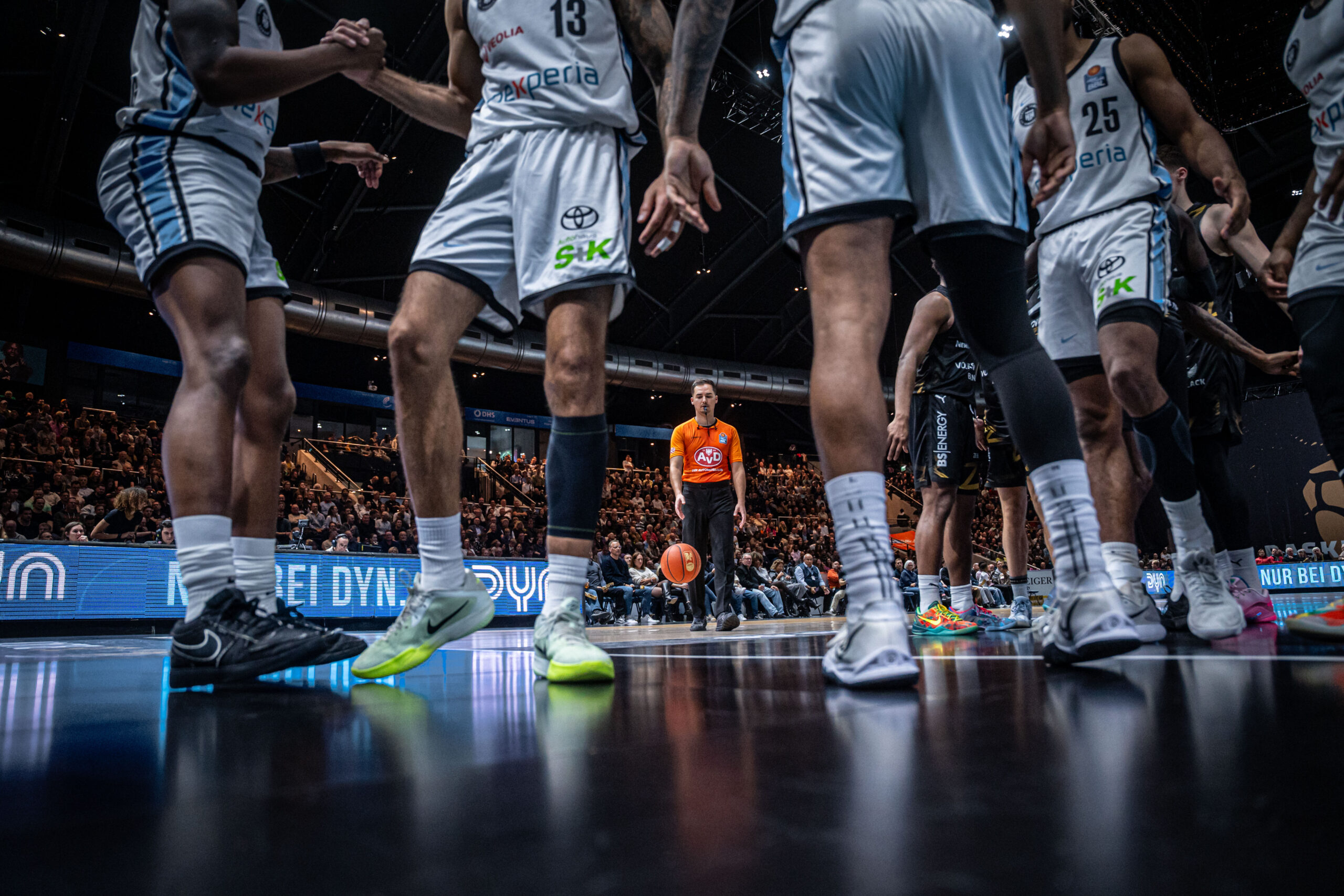 Mehrere Basketballspieler in weißen Trikots stehen auf dem Spielfeld, im Hintergrund ein Schiedsrichter in orangefarbenem Trikot und Zuschauer auf den Tribünen