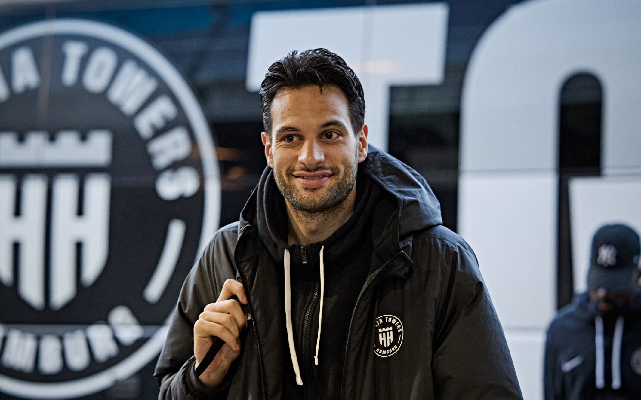 Mann mit schwarzer Jacke und Kapuzenpullover vor einem Fahrzeug mit dem Logo der Hamburg Towers