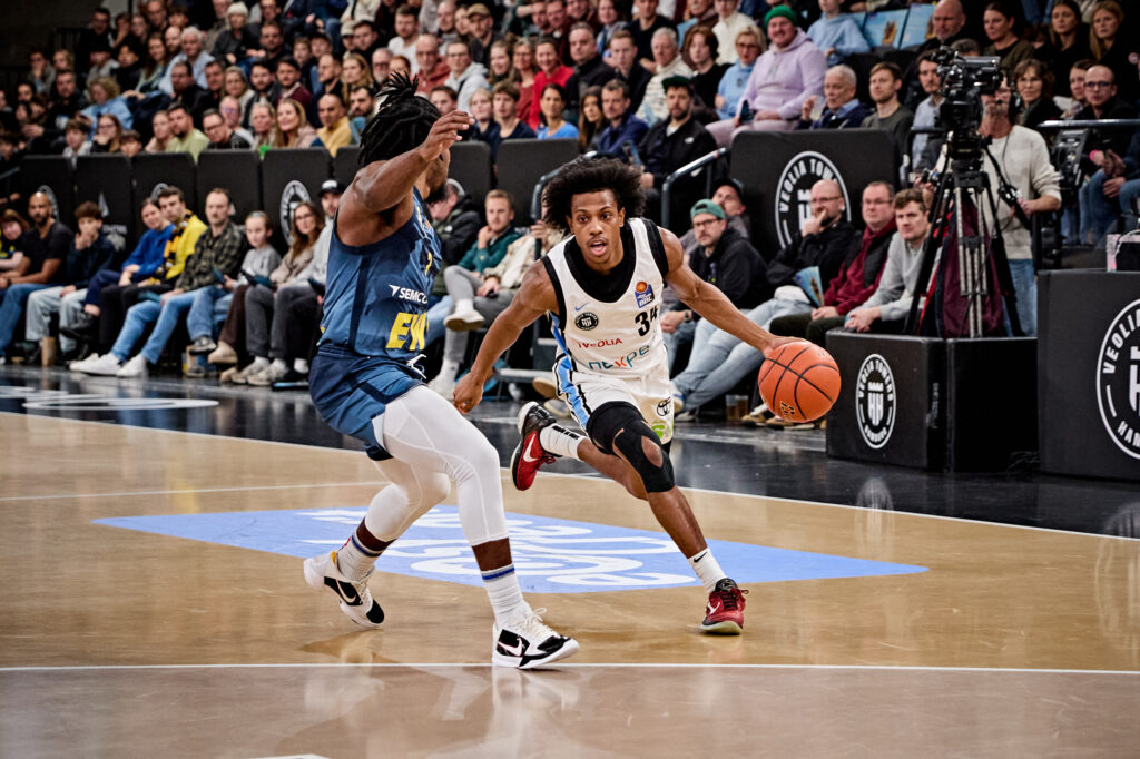 Zwei Basketballspieler in Aktion auf dem Spielfeld, einer dribbelt den Ball, der andere verteidigt, im Hintergrund Zuschauer und Kamerateam