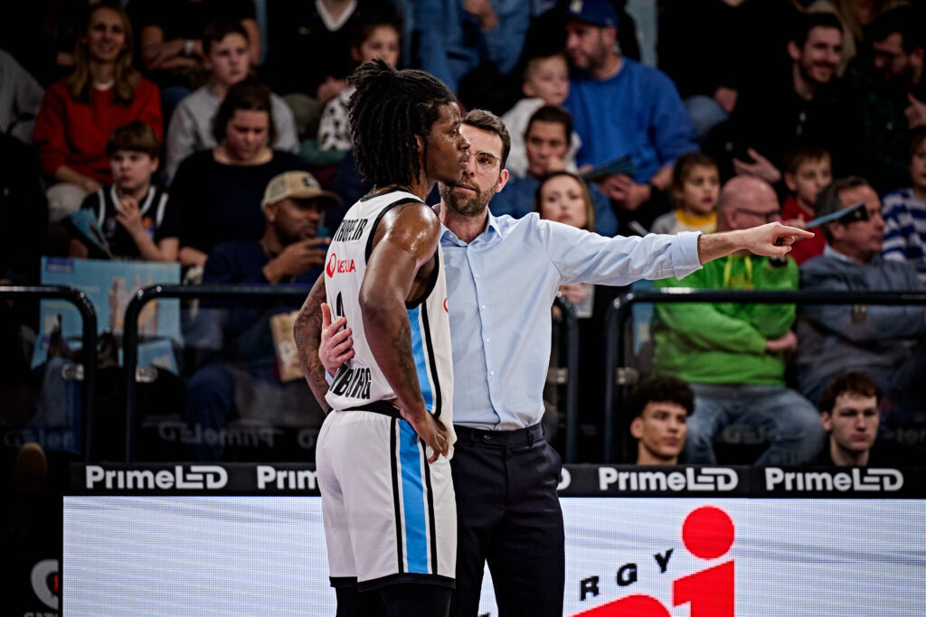 Ein Basketballspieler in weißem Trikot mit der Nummer 1 steht neben einem Mann in Hemd, der mit ausgestrecktem Arm in eine Richtung zeigt, im Hintergrund Zuschauer