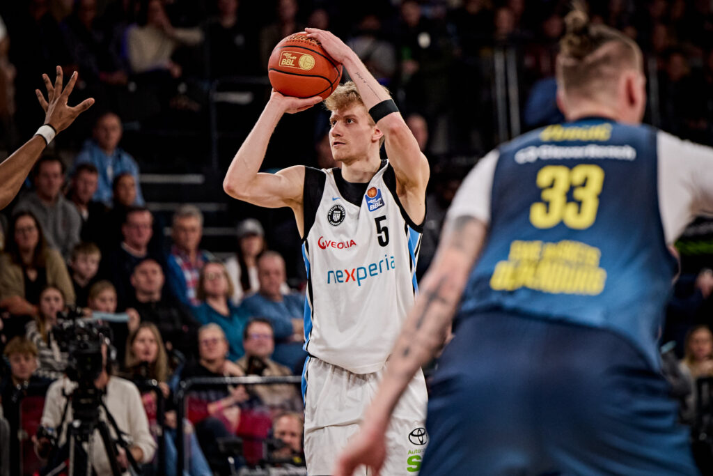 Basketballspieler in weißem Trikot mit der Nummer 5 hält den Ball über dem Kopf, während ein Spieler in blauem Trikot mit der Nummer 33 vor ihm steht