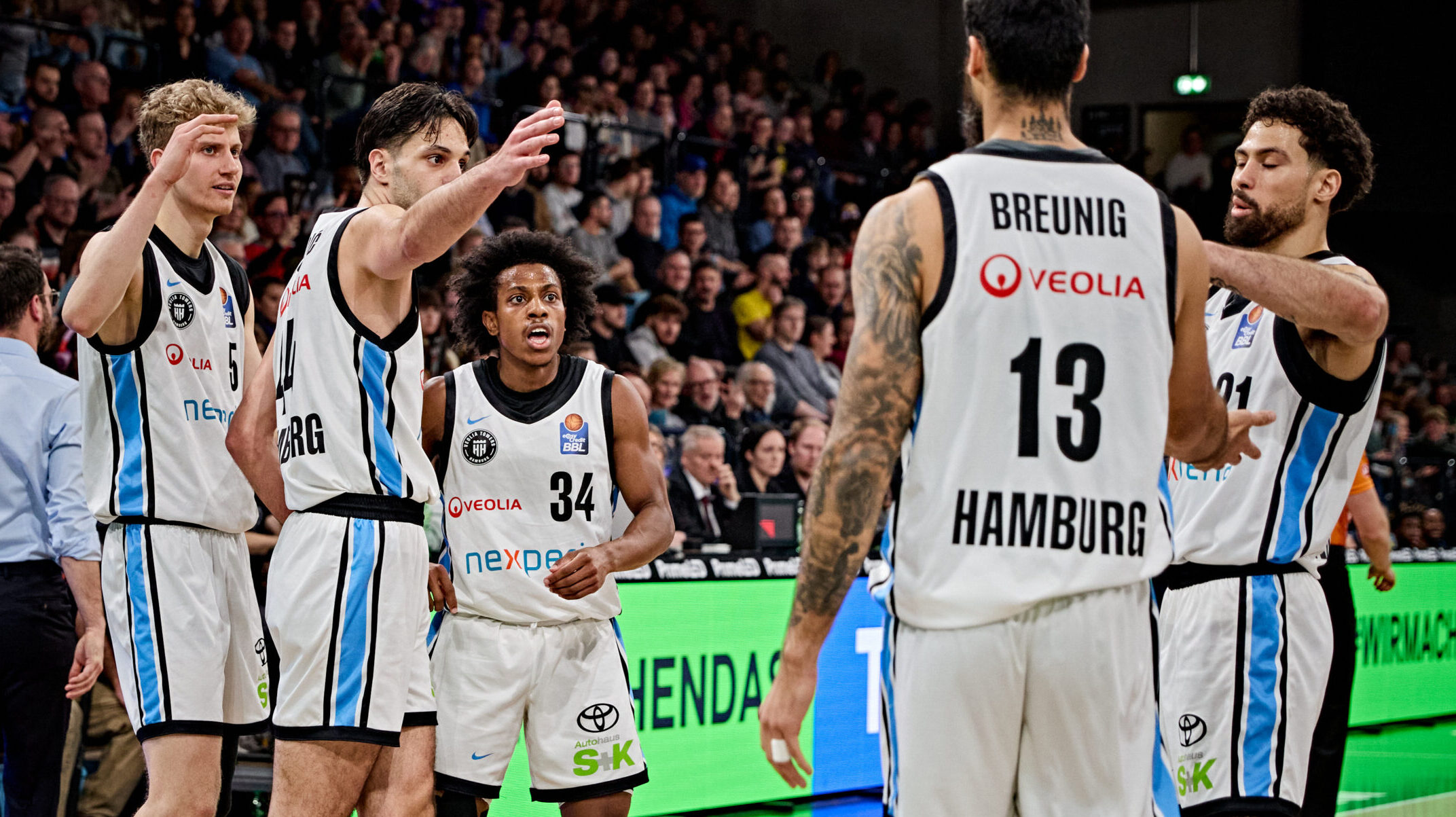 Mehrere Basketballspieler in weißen Trikots mit blauen und schwarzen Akzenten stehen auf dem Spielfeld, einer trägt die Rückennummer 13 mit dem Schriftzug 'BREUNIG HAMBURG'