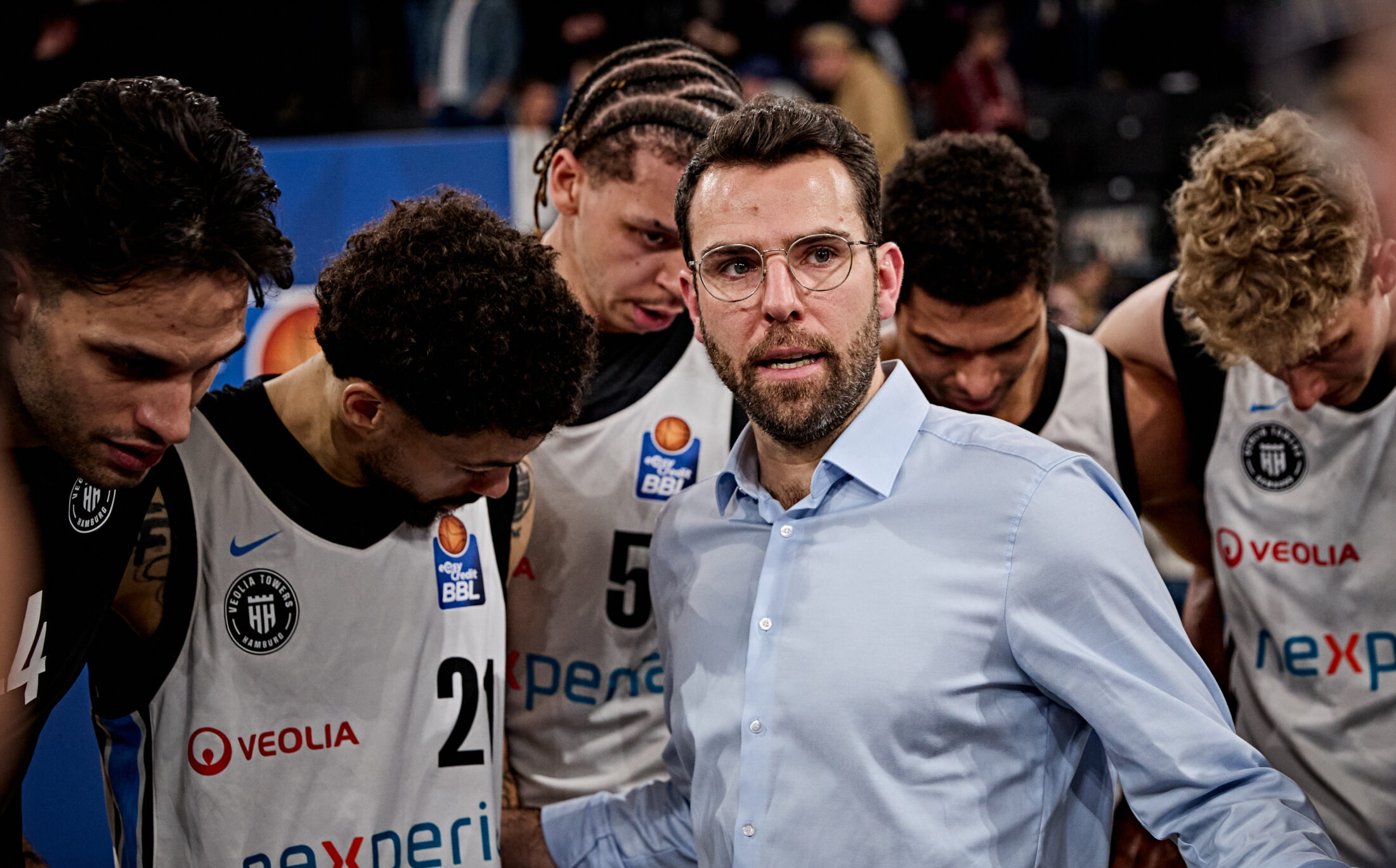Basketballspieler in Trikots mit den Logos von Veolia und Nexperia stehen um einen Mann in hellblauem Hemd in einer Sporthalle