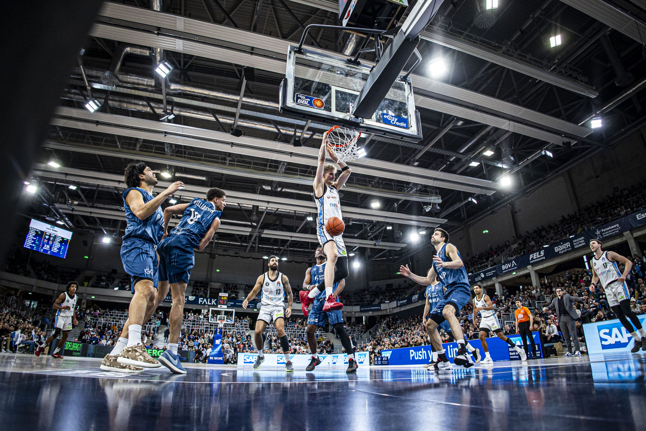 Mehrere Basketballspieler in Aktion unter dem Korb in einer Halle mit Zuschauern auf den Rängen.