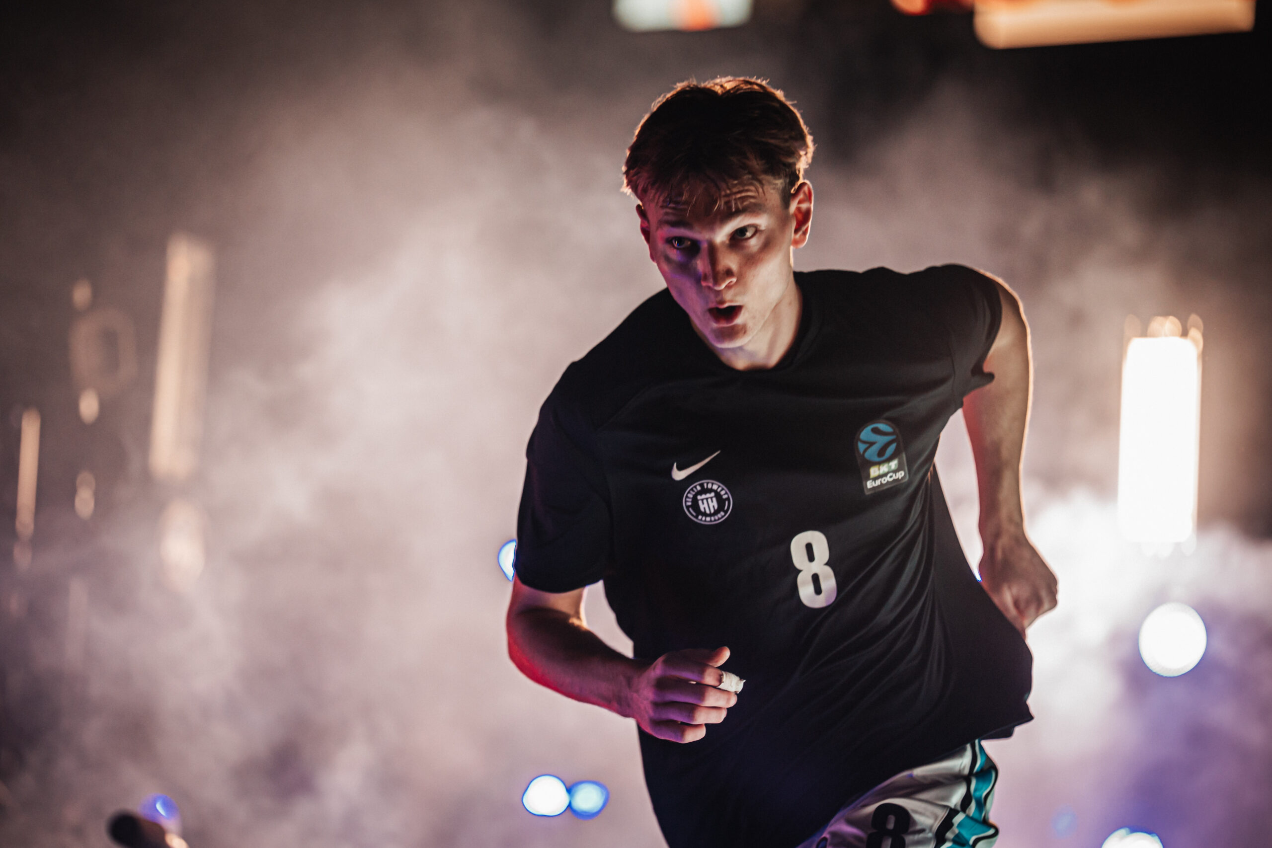 Junger Mann in schwarzem Sporttrikot mit der Nummer 8 läuft vor einem nebligen Hintergrund mit Lichtquellen
