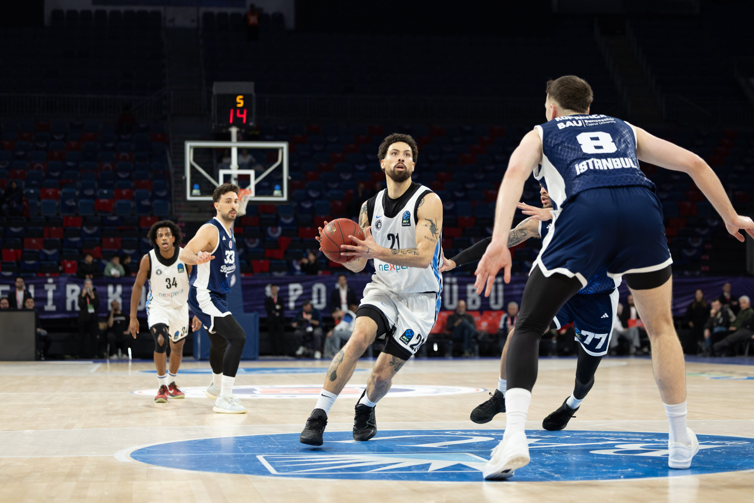 Mehrere Basketballspieler auf dem Spielfeld, einer in weißer Trikotnummer 24 hält den Ball und wird von einem Spieler in blauem Trikotnummer 8 verteidigt