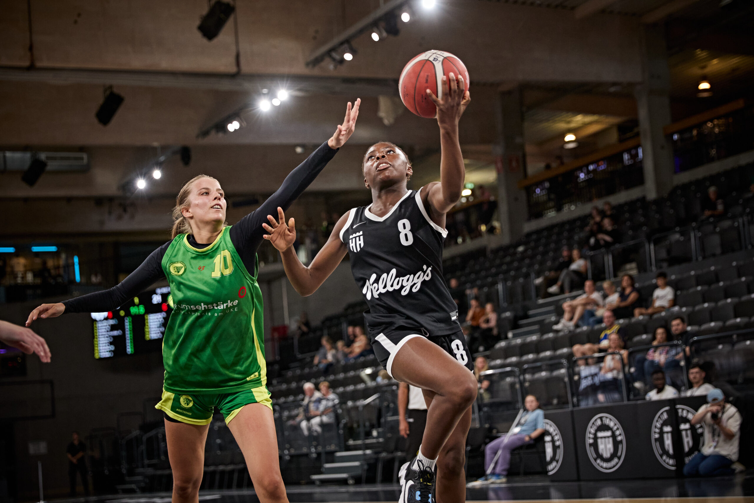 Basketballspiel mit einer Spielerin in schwarzem Trikot mit Ball in der Luft und einer Spielerin in grünem Trikot, die verteidigt