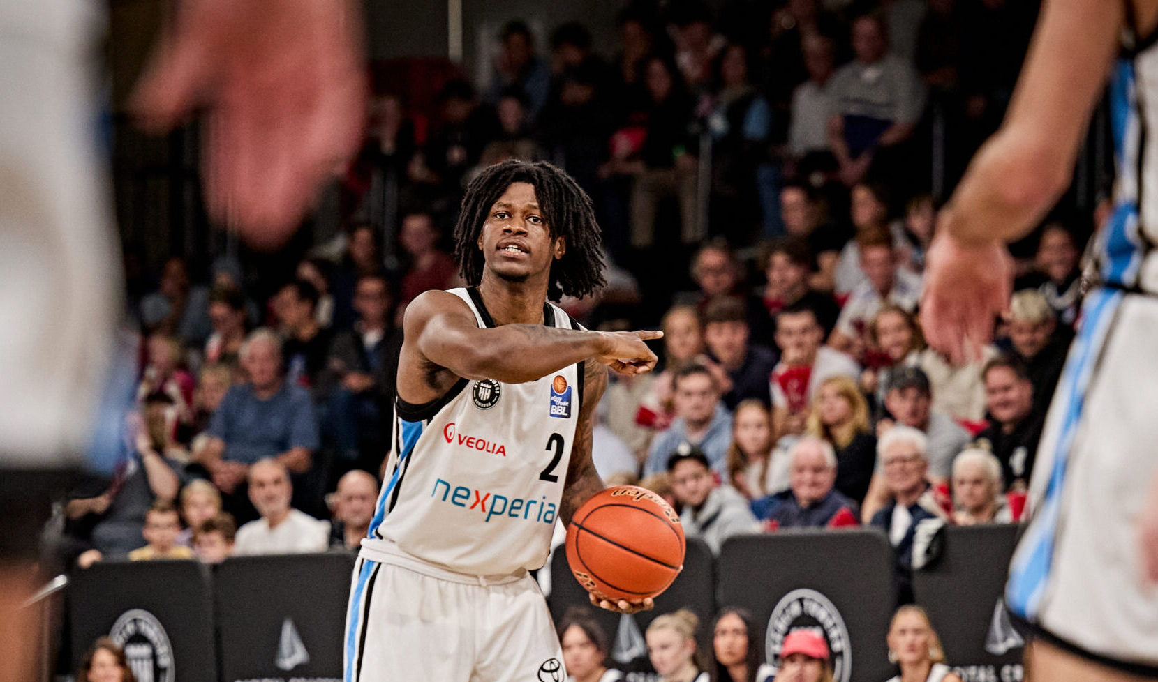Basketballspieler mit Trikotnummer 2 passt den Ball zu einem Mitspieler auf dem Spielfeld in einer Halle mit Zuschauern im Hintergrund