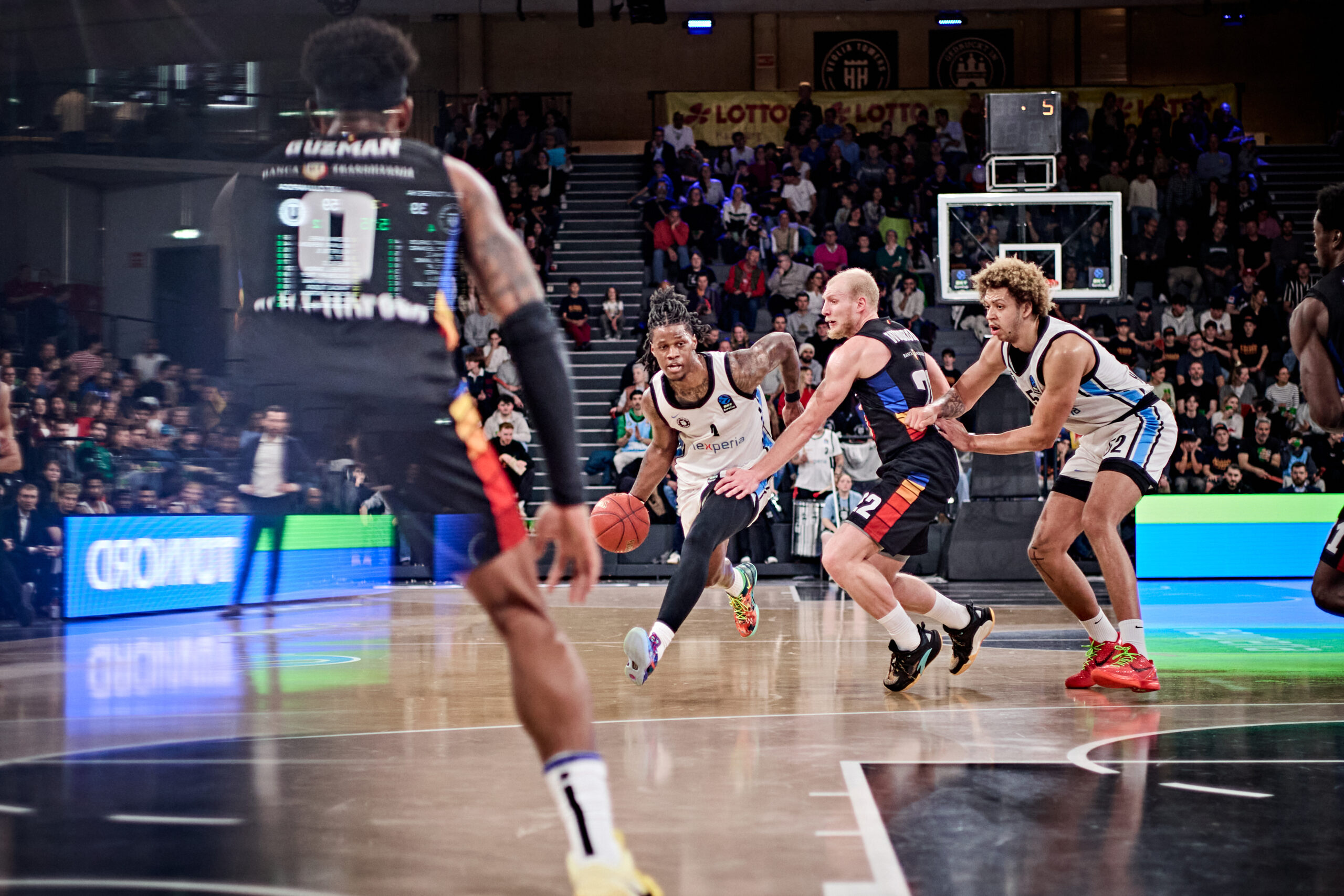 Mehrere Basketballspieler in Aktion auf dem Spielfeld, einer dribbelt den Ball, andere verteidigen ihn.
