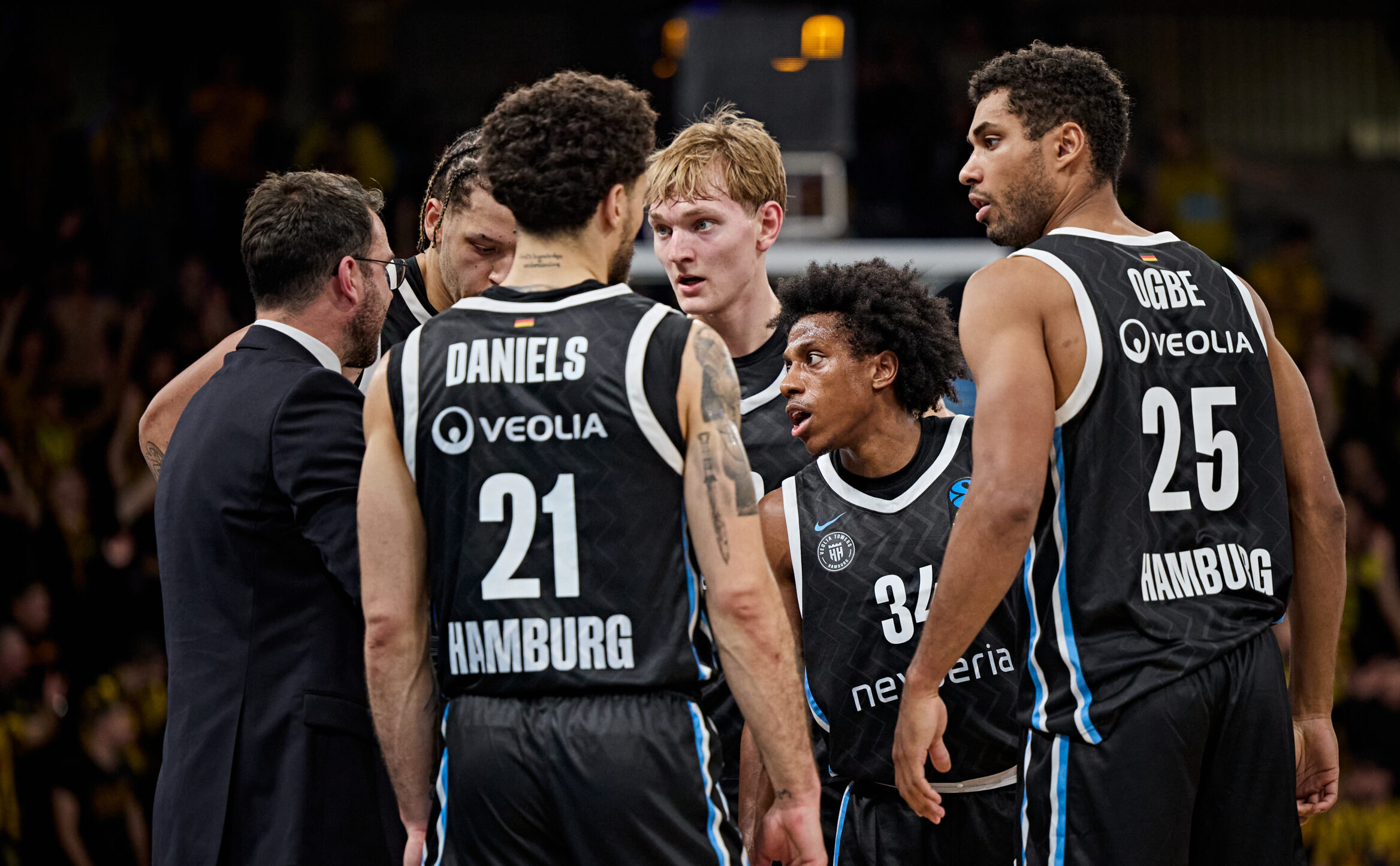 Mehrere Basketballspieler in schwarzen Trikots mit den Aufschriften 'DANIELS 21 HAMBURG' und 'OGBE 25 HAMBURG' stehen in einer Gruppe mit einem Trainer in Anzug in einer Halle.