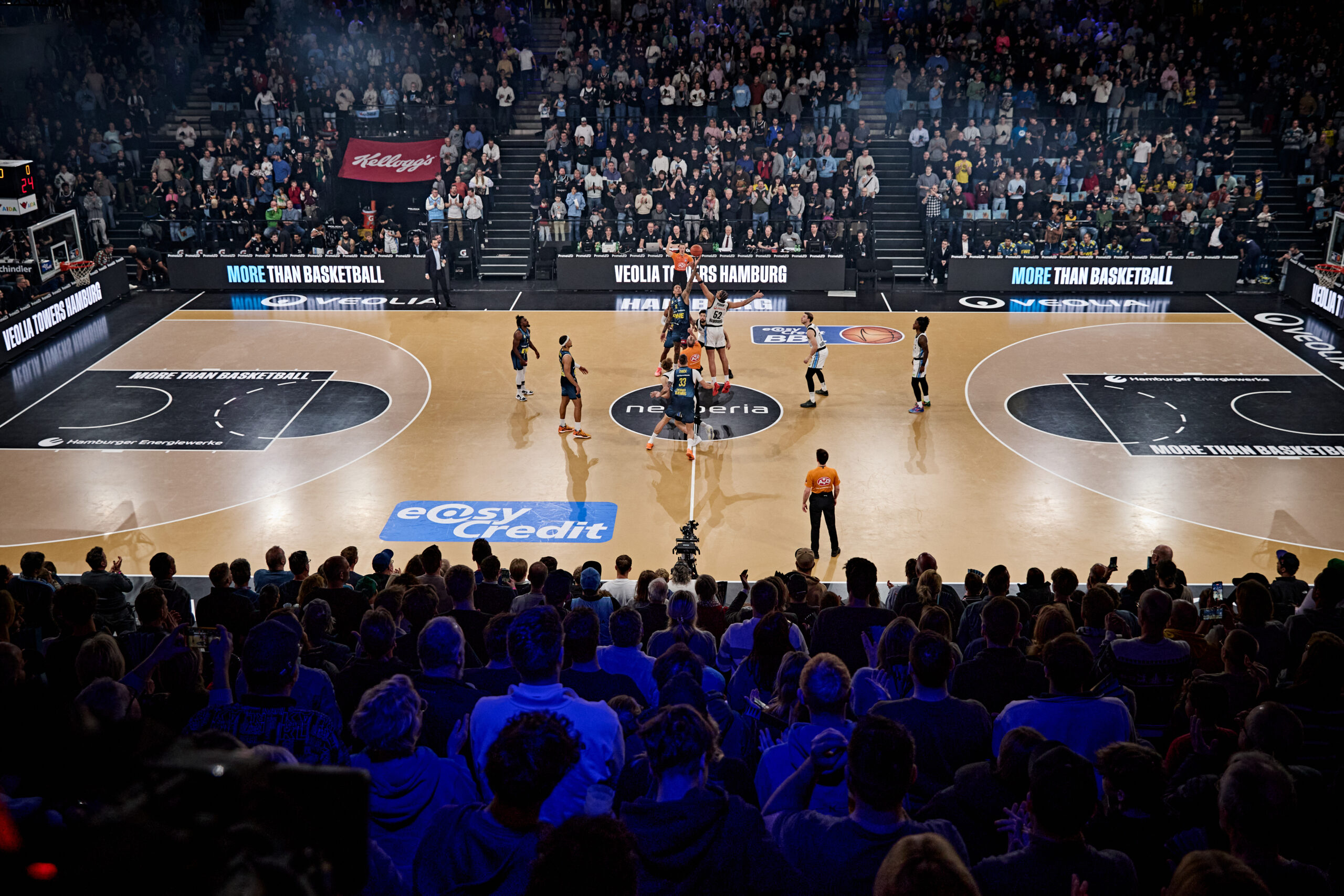Basketballspiel mit Spielern beim Sprungball auf einem Spielfeld vor voller Zuschauerkulisse in einer Halle