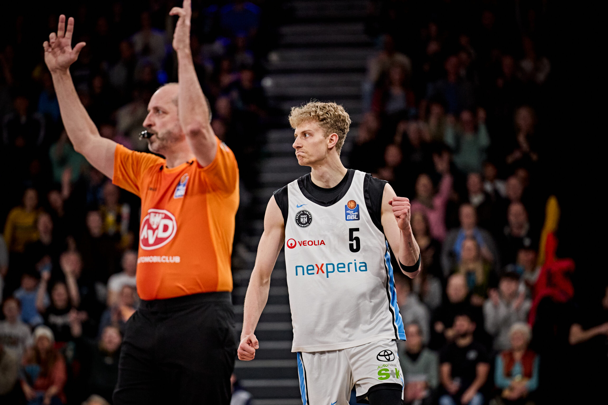 Basketballspieler in weißem Trikot mit der Nummer 5 und erhobener Faust auf dem Spielfeld, im Hintergrund ein Schiedsrichter in orangefarbenem Trikot mit erhobenen Armen