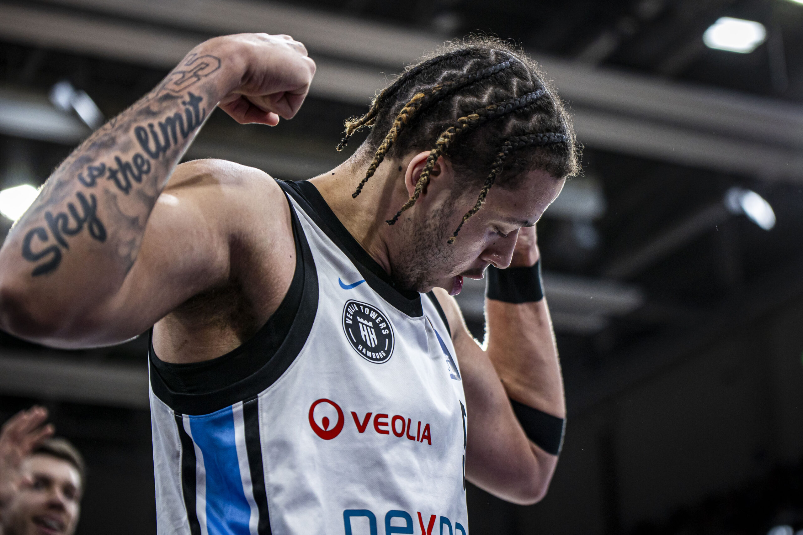 Mann mit geflochtenem Haar und Muskelarm zeigt tätowierten Oberarm mit Schriftzug 'Sky the limit' und trägt ein Trikot mit den Logos 'VEOLIA' und 'nexpa'