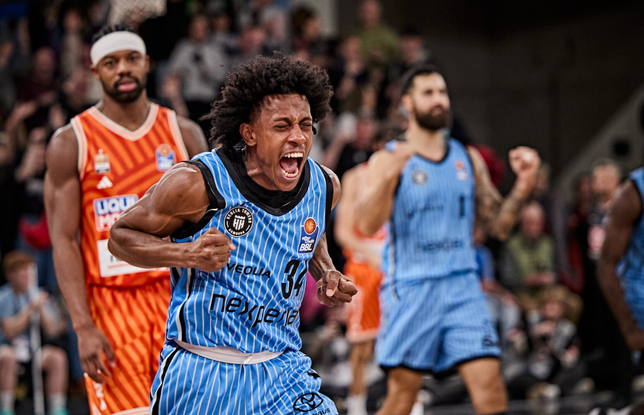 Basketballspieler in blau-weiß gestreiftem Trikot mit der Nummer 34 in Aktion vor Spielern in orangefarbenen und hellblauen Trikots in einer Halle mit Publikum im Hintergrund