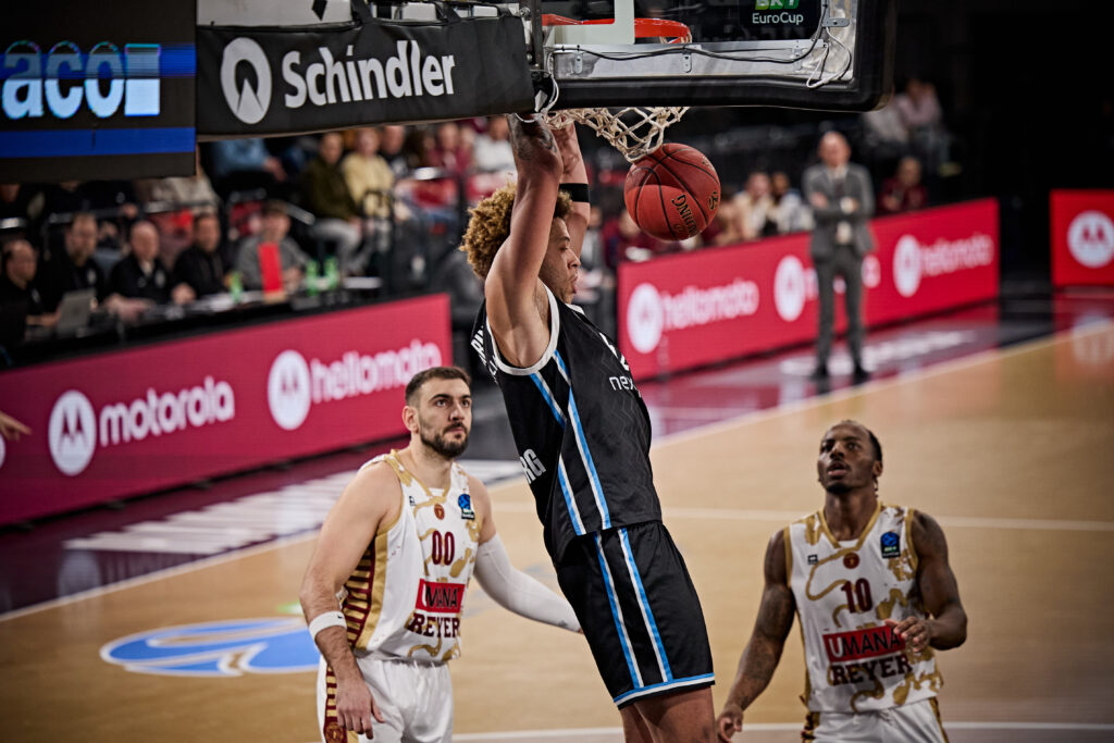 Basketballspieler in schwarzem Trikot hängt am Korb und führt einen Dunk aus, zwei Spieler in weißen Trikots beobachten das Spiel.