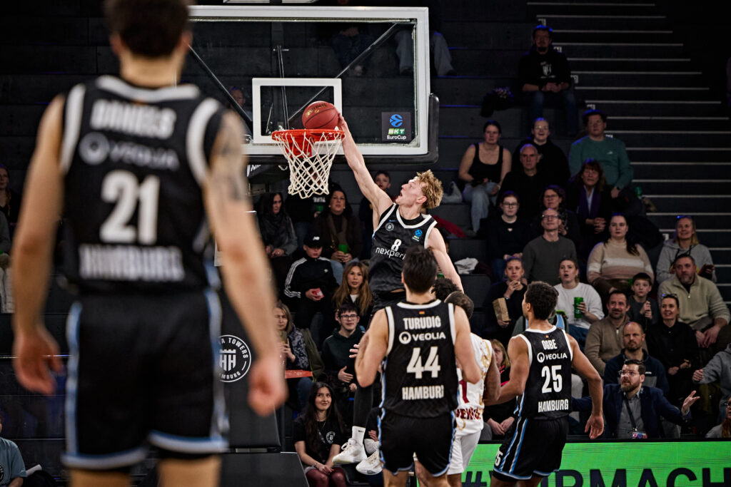 Basketballspieler in schwarzen Trikots der Hamburg Towers beim Korbleger vor dem Korb in einer Halle mit Zuschauern im Hintergrund
