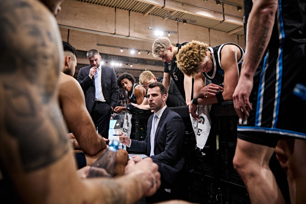 Basketballtrainer im Anzug gibt Anweisungen an Spieler in schwarzen Trikots während einer Auszeit in der Halle