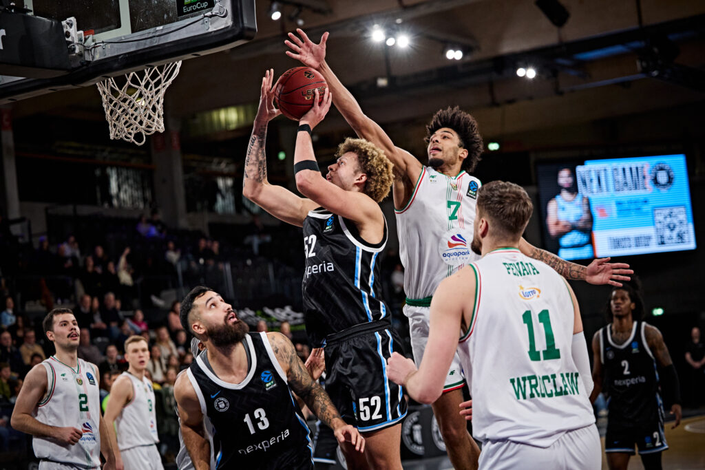 Mehrere Basketballspieler kämpfen um den Ball unter dem Korb in einer Halle während eines Spiels.