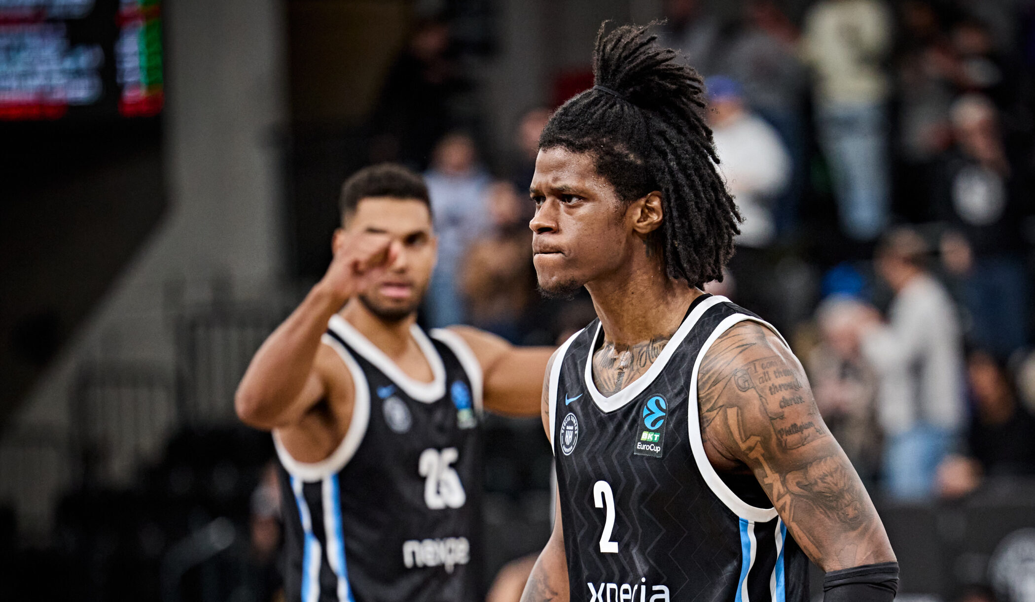Zwei Basketballspieler in schwarzen Trikots mit der Aufschrift 'Nexperia' auf dem Spielfeld, einer mit erhobenen Armen, der andere mit Dreadlocks und sichtbaren Tattoos am rechten Arm.