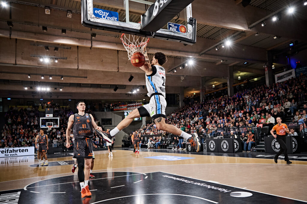 Ein Basketballspieler in weiß-schwarzer Uniform springt zum Dunking in einen vollen Halleninnenraum mit Zuschauern.