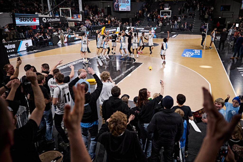 Basketballspieler auf dem Spielfeld und jubelnde Zuschauer am Spielfeldrand in einer Halle