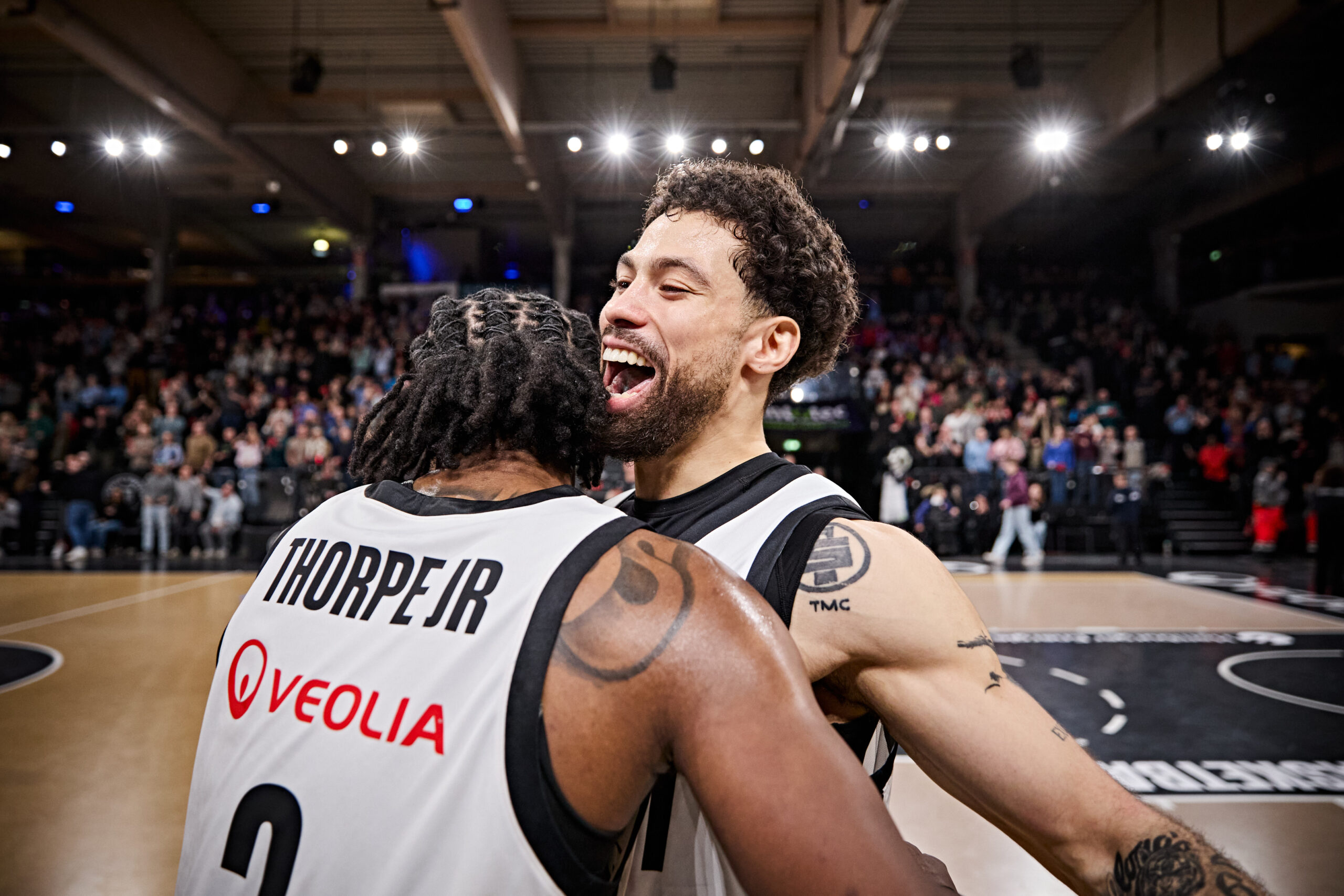 Zwei Basketballspieler in weißen Trikots umarmen sich auf dem Spielfeld vor voller Zuschauerkulisse