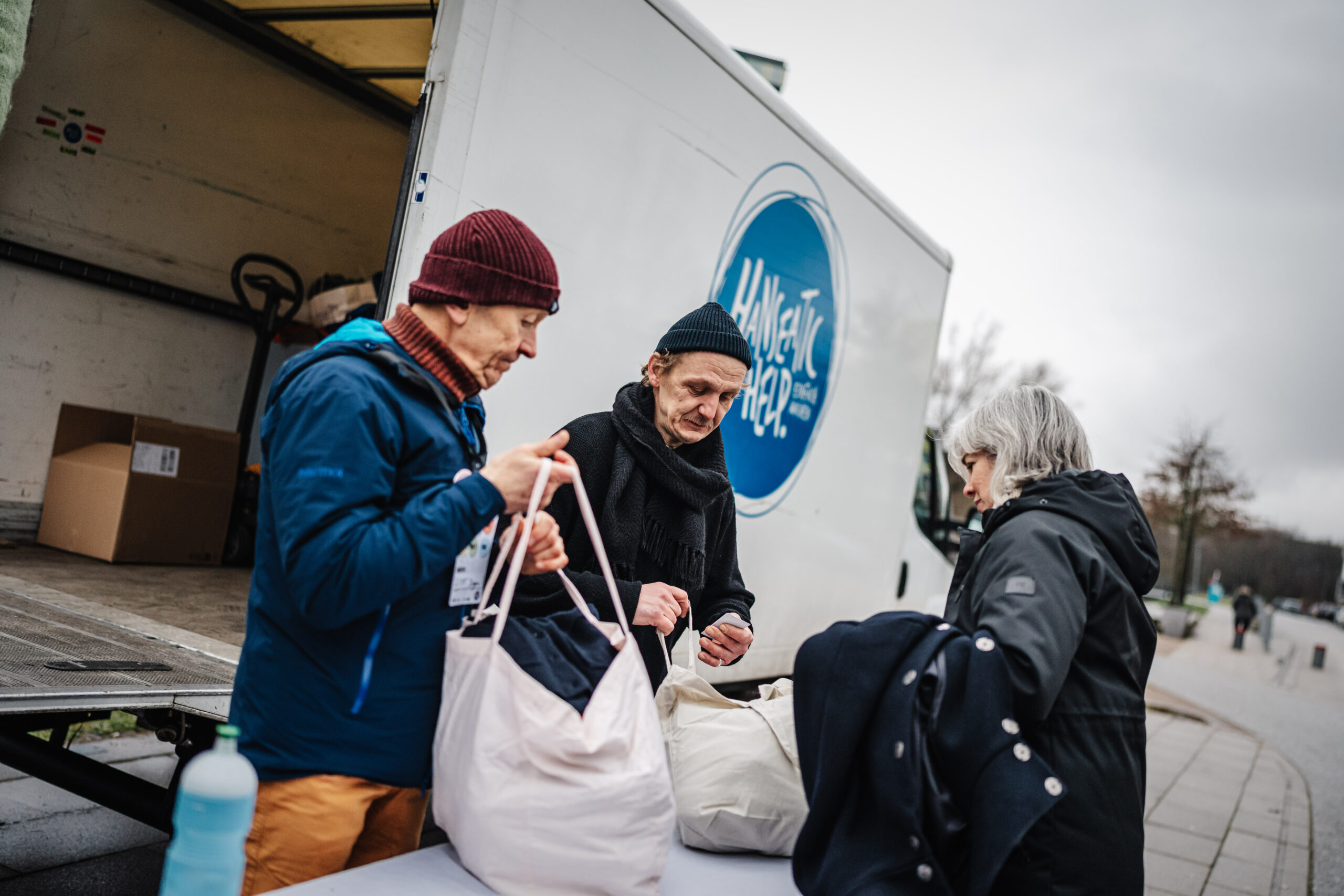Drei Personen mit Winterkleidung stehen vor einem weißen Lieferwagen mit dem blauen Logo "Hanseatic Help" und übergeben weiße Taschen.