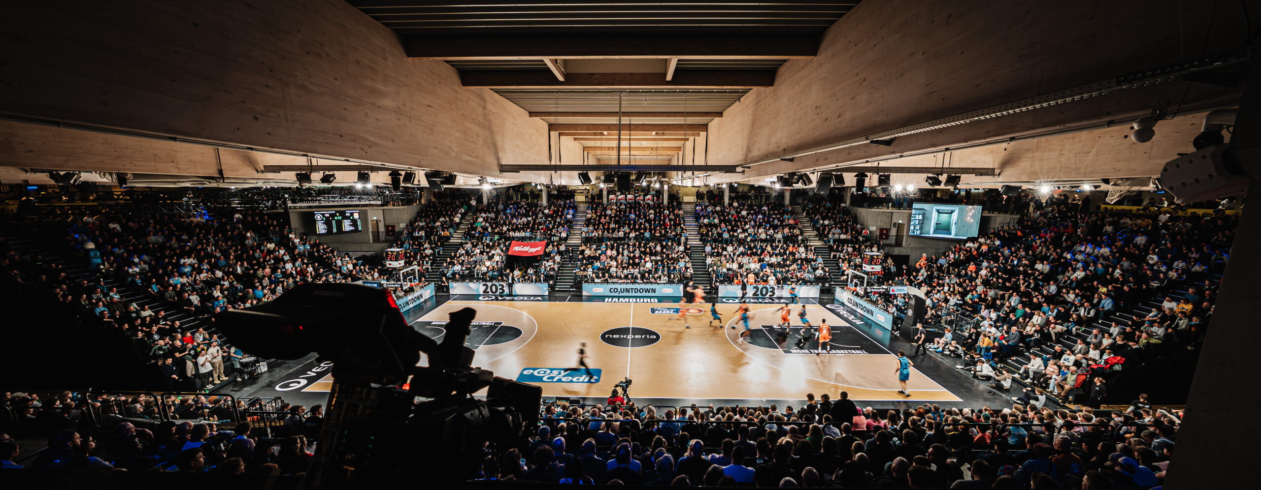 Weitwinkelaufnahme einer vollbesetzten Basketballhalle mit laufendem Spiel auf dem Spielfeld