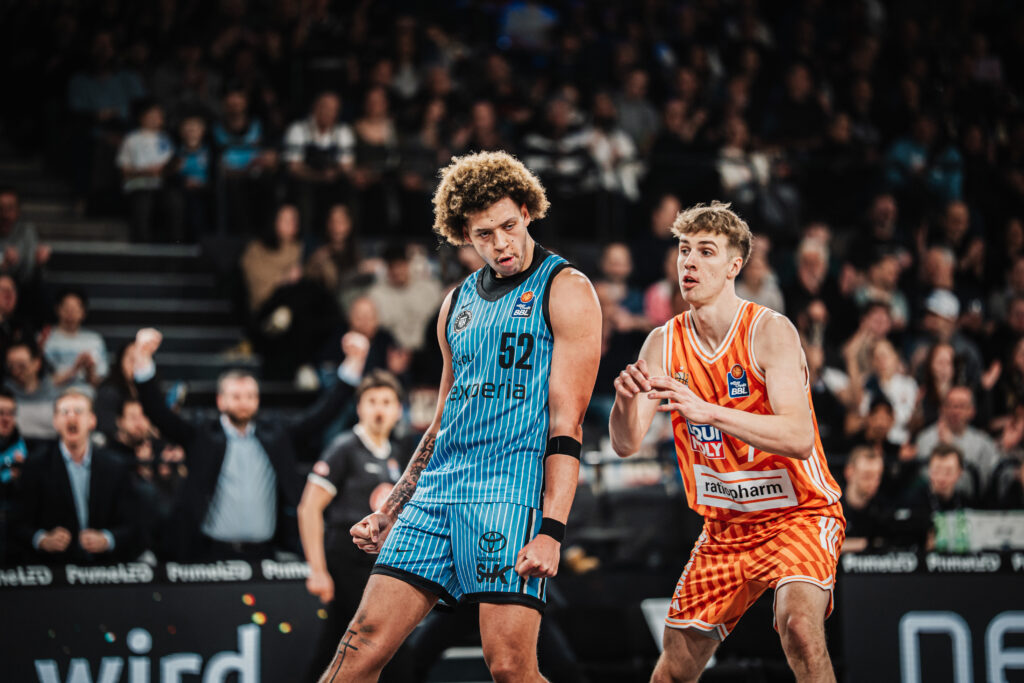 Zwei Basketballspieler in Aktion auf dem Spielfeld, einer in blauem Trikot mit der Nummer 52, der andere in orange-weiß kariertem Trikot
