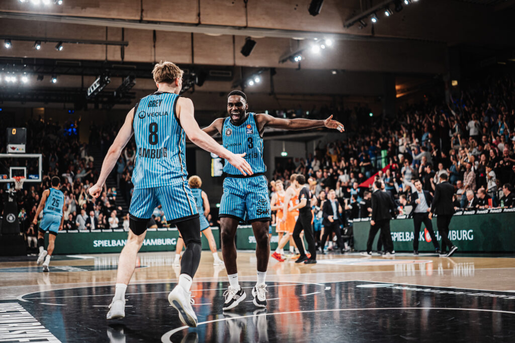 Zwei Basketballspieler in gestreiften Trikots mit den Nummern 8 und 3 auf dem Spielfeld vor jubelndem Publikum