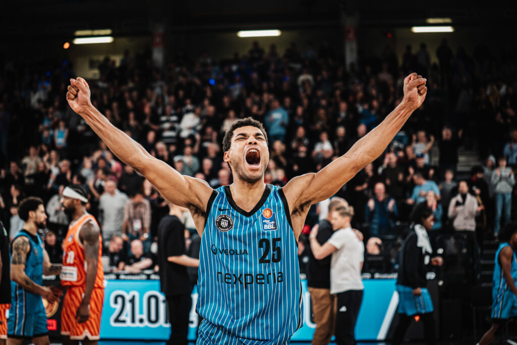 Basketballspieler mit hochgereckten Armen in blau-schwarzem Trikot mit der Nummer 25 vor Publikum in einer Halle