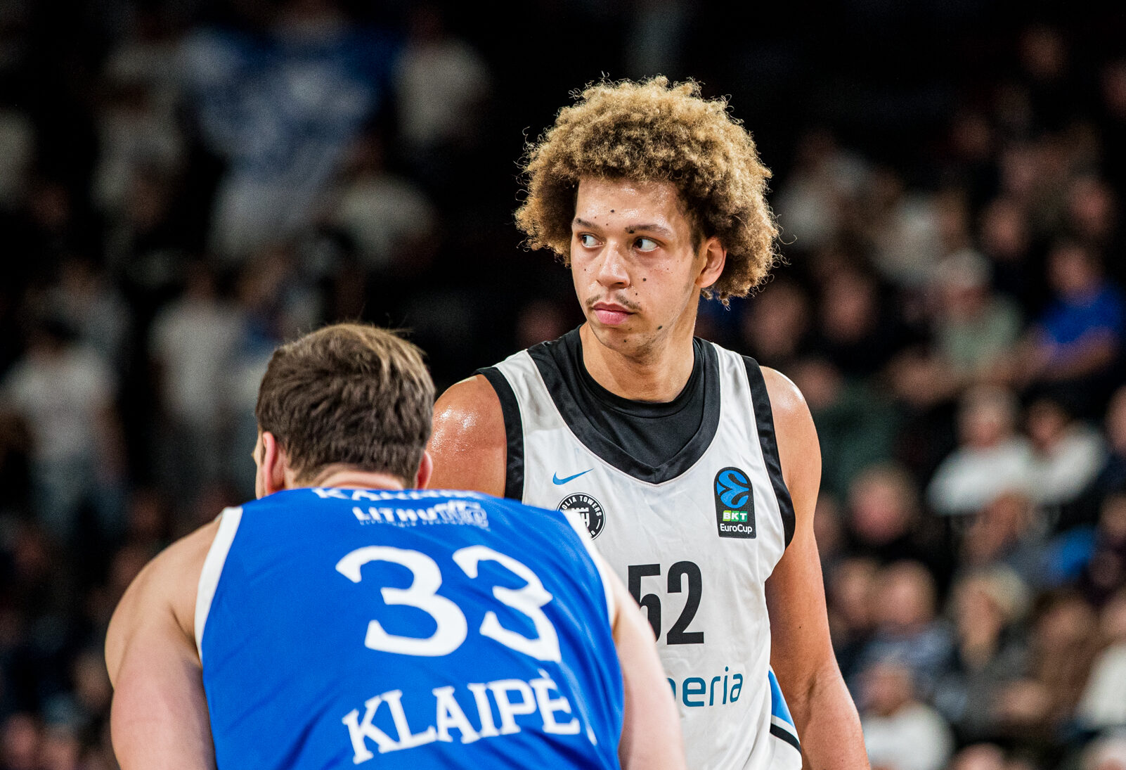 Zwei Basketballspieler, einer in weißem Trikot mit der Nummer 52, der andere in blauem Trikot mit der Nummer 33 und der Aufschrift "KLAIPE", vor einem Publikum in einer Halle.