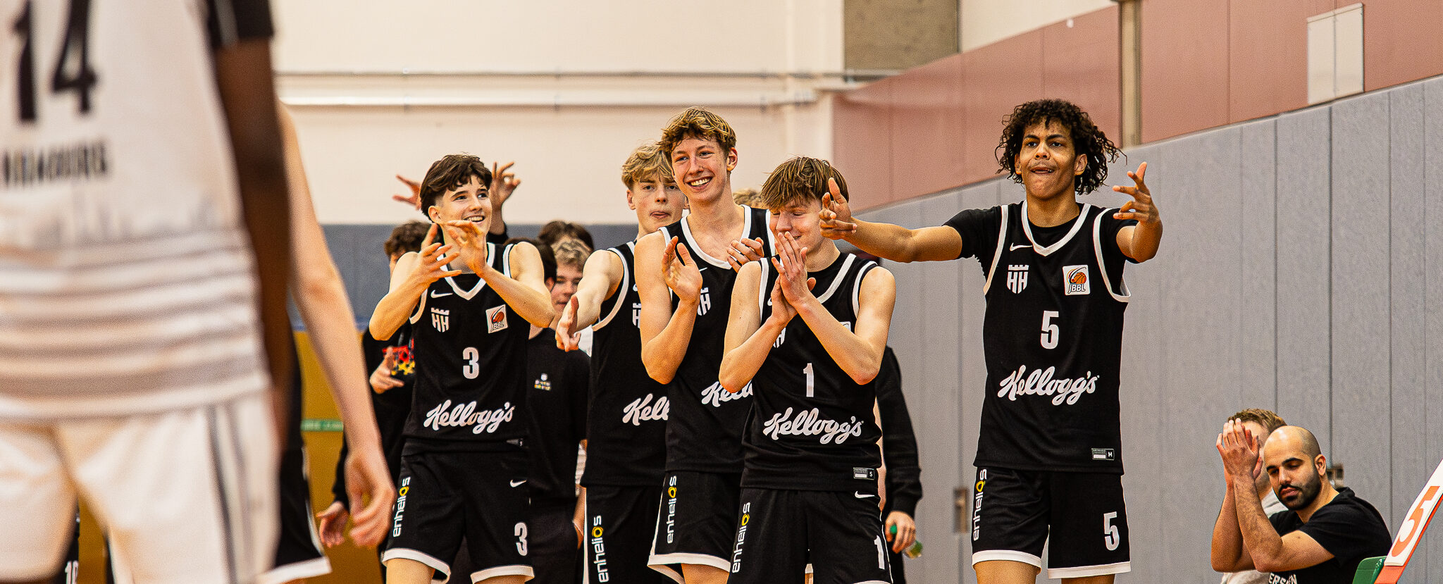 Fünf Basketballspieler in schwarzen Trikots mit der Aufschrift "Kellogg's" klatschen und gestikulieren in einer Halle.
