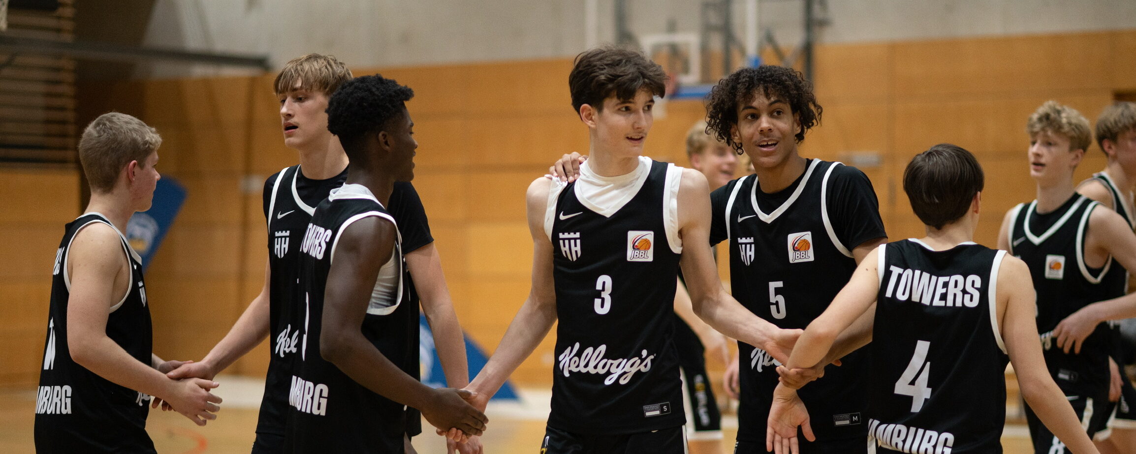 Jugendliche Basketballspieler in schwarzen Trikots stehen in einer Halle im Kreis und halten sich an den Händen.