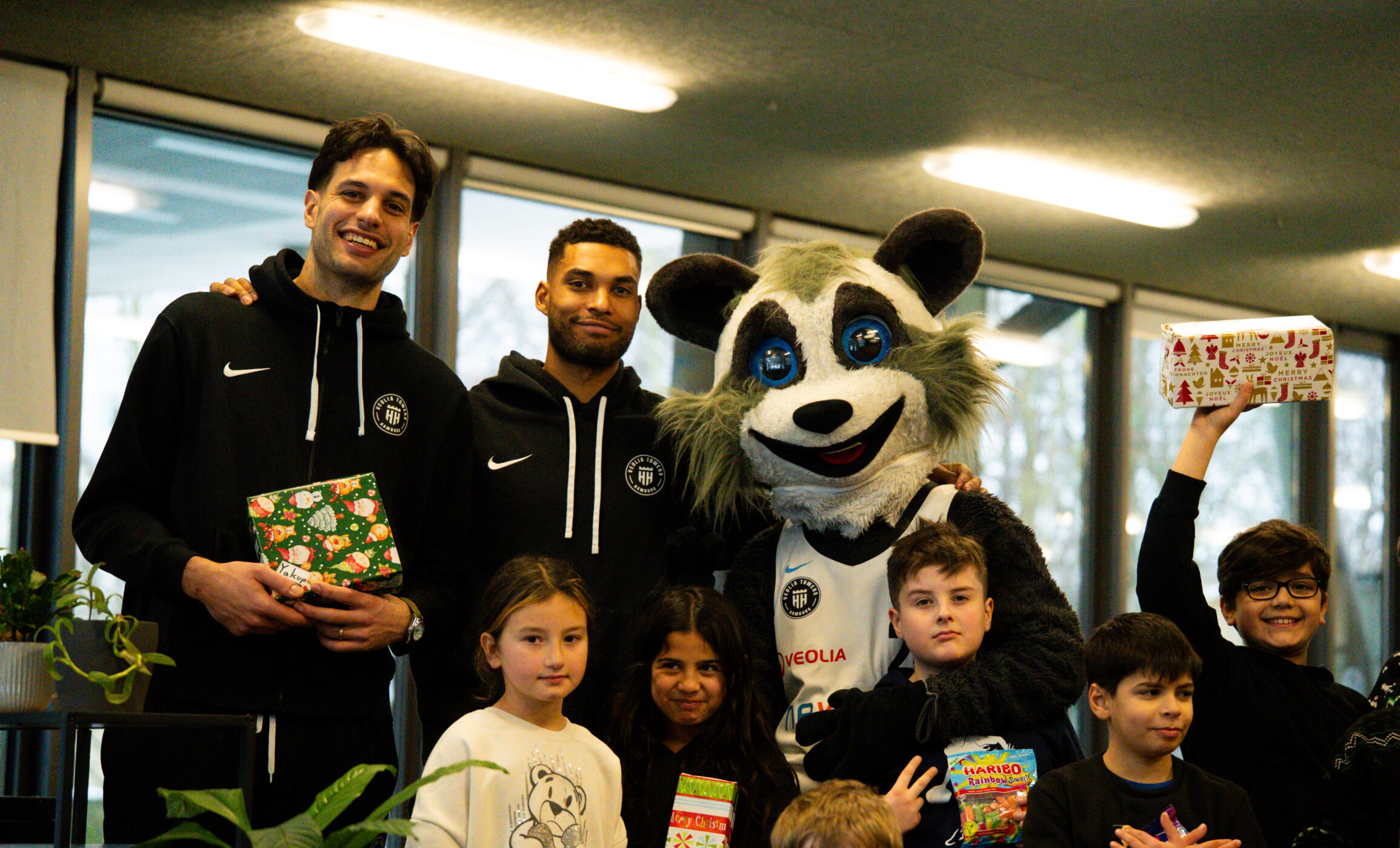 Gruppenfoto mit zwei Personen in schwarzen Sportjacken, einem Maskottchen mit Tierkostüm und mehreren Kindern, die Geschenke halten.