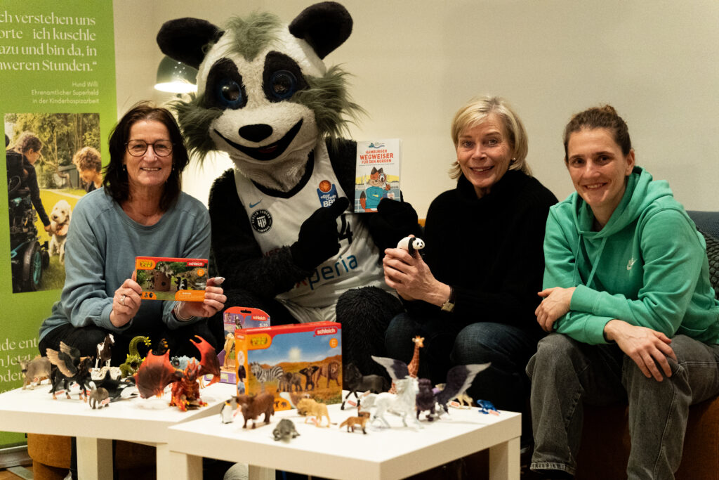 Vier Personen sitzen um einen Tisch mit vielen Tierfiguren und zwei Spielzeugverpackungen, in der Mitte eine Person im Waschbärkostüm mit Daumen hoch